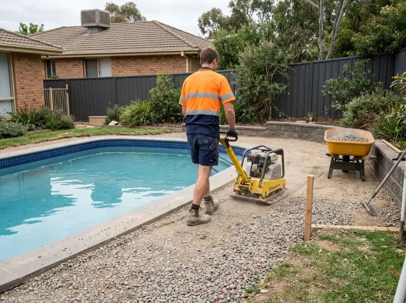 Worker running a plate compactor over crushed rock road base laid around the edge of a pool