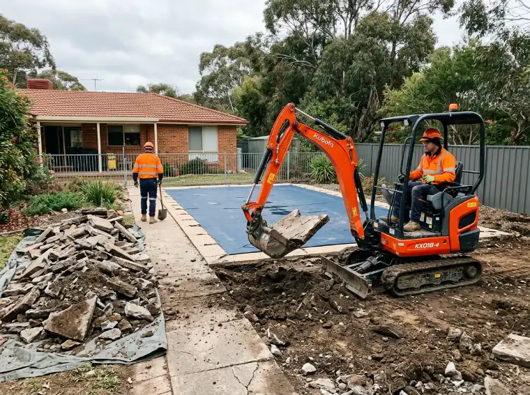 Mini excavator lifting old concrete pool paving into a pile while the pool sits under a cover